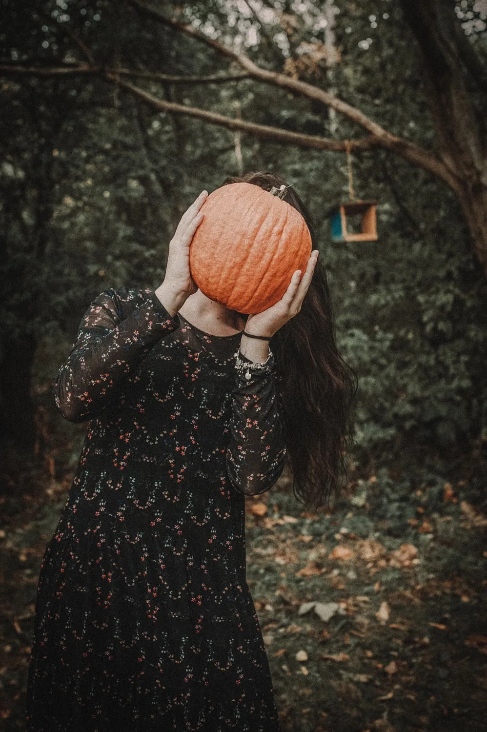 Witchy outfit ideas with a black floral dress and orange pumpkin in a forest
