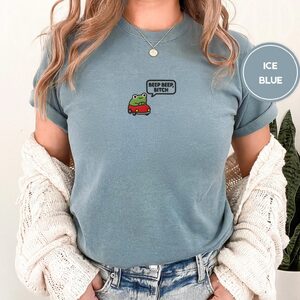 Person wearing an ice blue Beep Beep Driving Frog Embroidered T-Shirt