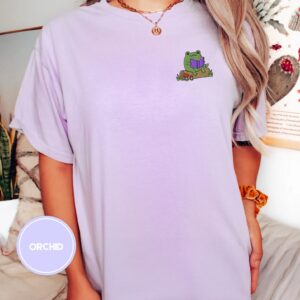 Person wearing an orchid Frog Reading On A Log Embroidered T-Shirt