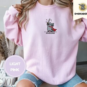 Person wearing a light pink Huzzah Raccoon Knight Embroidered Sweatshirt