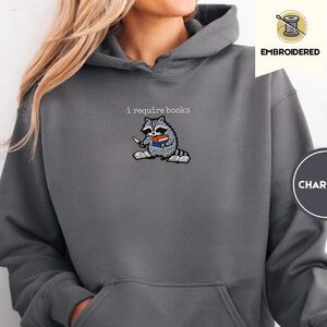 Person wearing a charcoal I Require Books Raccoon Embroidered Hoodie