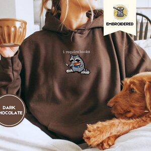 Person wearing a dark chocolate I Require Books Raccoon Embroidered Hoodie