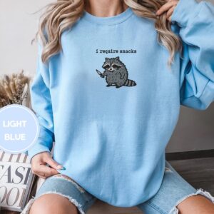 Person wearing a light blue I Require Snacks Raccoon Embroidered Sweatshirt