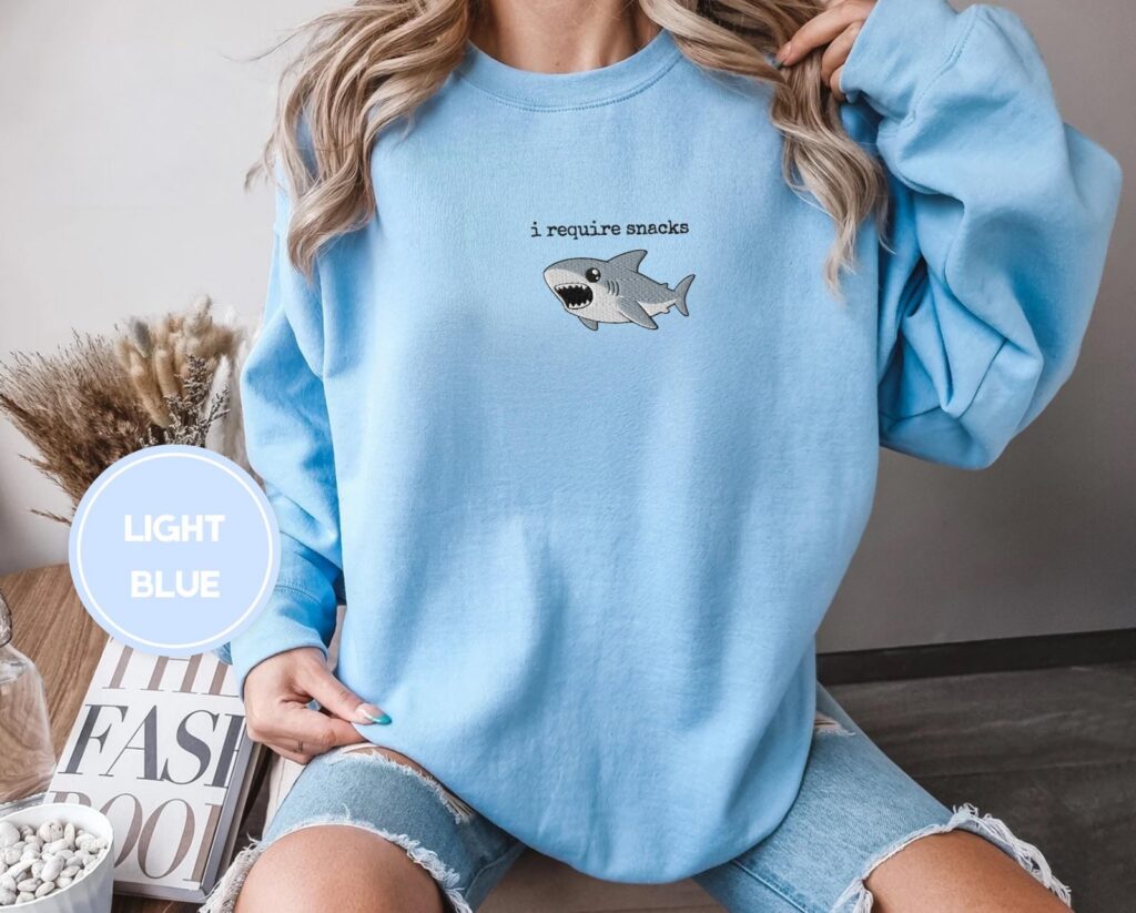Person wearing a light blue I Require Snacks Shark Embroidered Sweatshirt