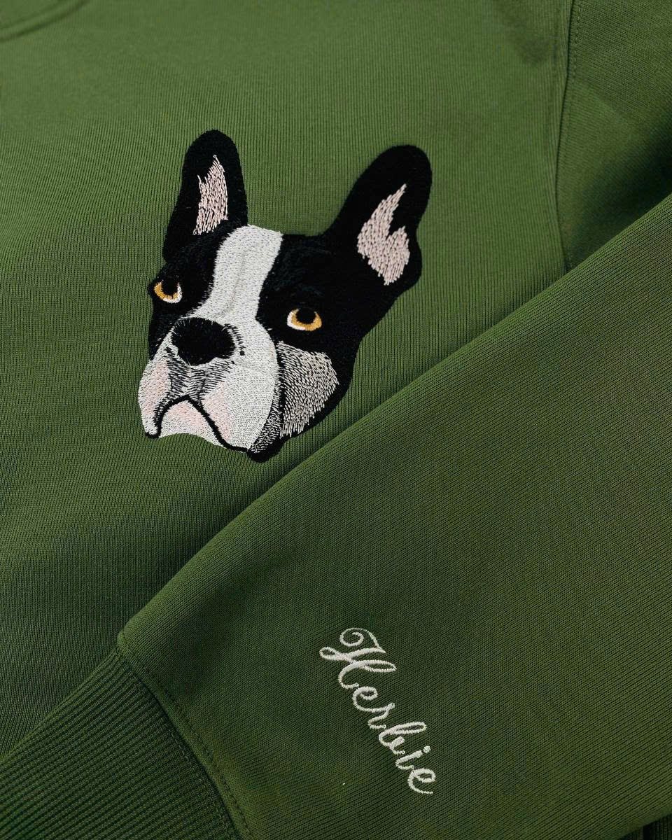 Close-up of a stitched French bulldog portrait on a green sweatshirt, with the name Herbie embroidered on the sleeve