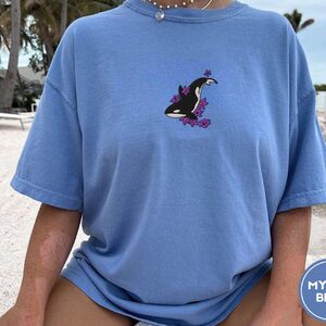 Person wearing a mystic blue Orca and Lilacs Embroidered T-Shirt