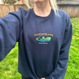 Person wearing a navy San Juan Islands embroidered sweatshirt with an orca tail and sailboat design