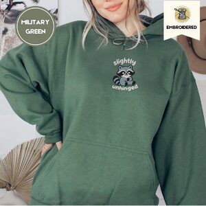 Person wearing a military green Slightly Unhinged Raccoon Embroidered Hoodie